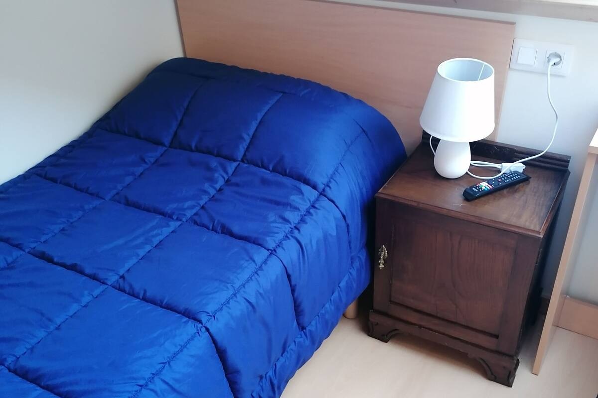 Un dormitorio con colcha azul, una mesita de noche de madera con lámpara y control remoto, y techo de madera.