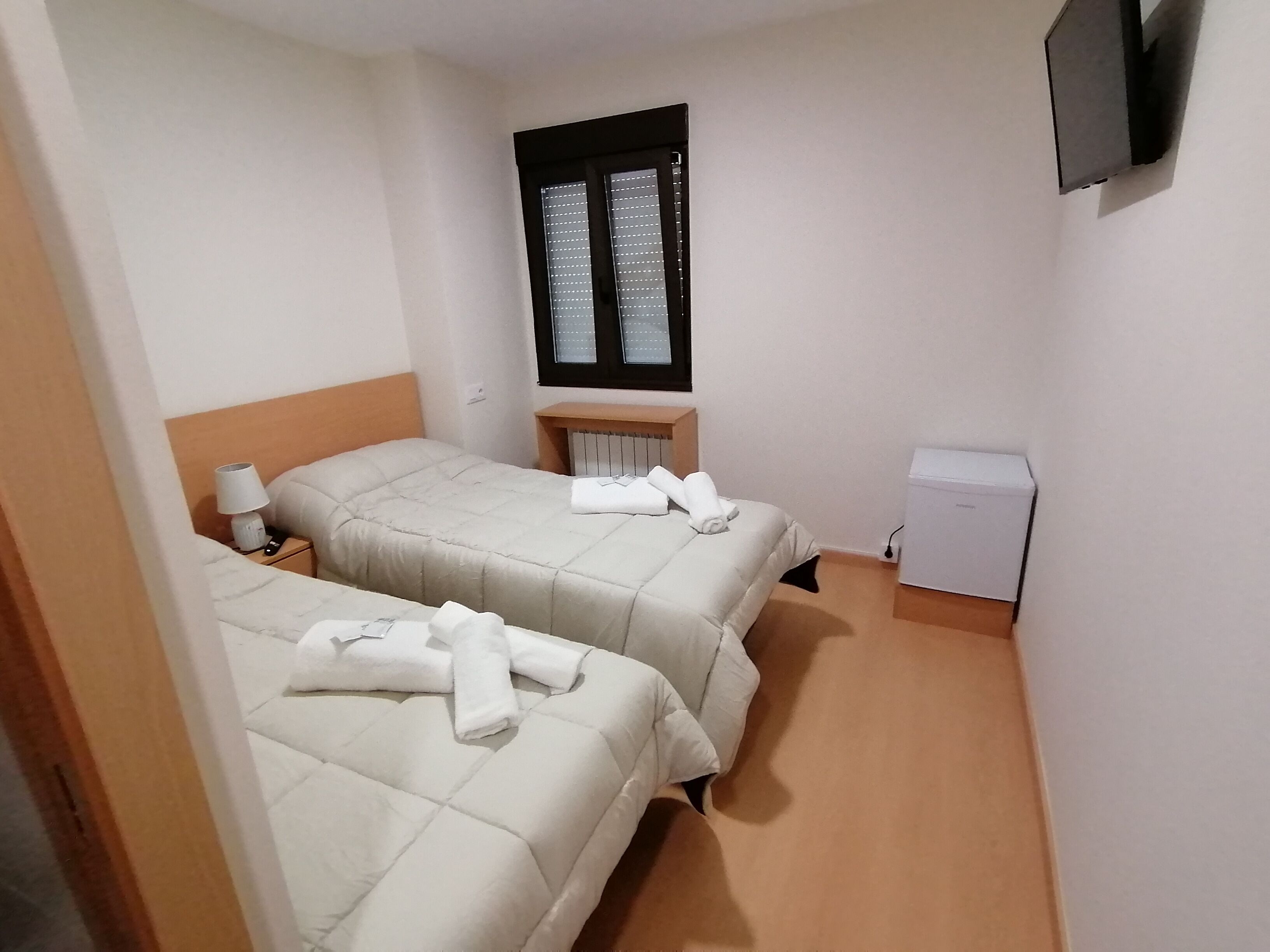 A small, clean room with two beds, a window, a radiator, a TV, and a small white cabinet.