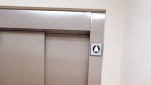 Puerta metálica de ascensor con panel de control que incluye una flecha direccional y un botón circular.