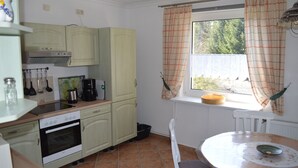 Eine Küche mit Herd, Backofen und Spüle, ein Esstisch mit Stühlen und ein Fenster mit Vorhängen.