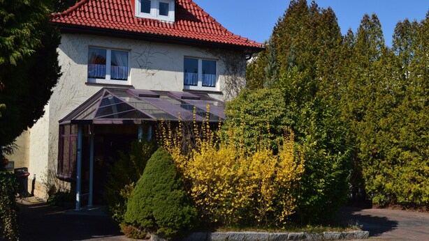 Ein zweigeschossiges Haus mit rotem Ziegeldach, einer Veranda und einem Garten mit gelb blühenden Büschen.