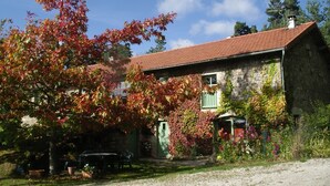 Une maison de campagne en pierre, avec un toit recouvert de tuiles rouges, une porte verte et un jardin où poussent diverses plantes et fleurs.