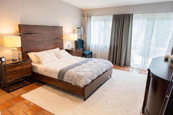 Une chambre d’hôtel avec un lit en bois, une table de chevet avec une lampe, une chaise, un bureau avec une lampe et une grande fenêtre avec des rideaux.
