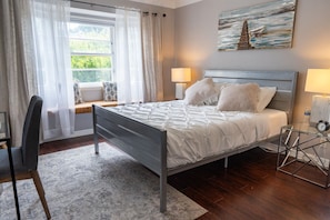 A bedroom with a bed, bedside lamps, a window with curtains, a chair, a small table, and a painting on the wall.