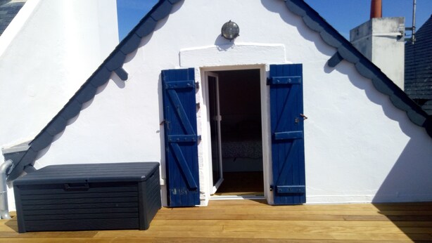 Een klein, afgesloten buitengebied met een houten terras, een zwarte opbergkist en een deur met blauwe luiken.