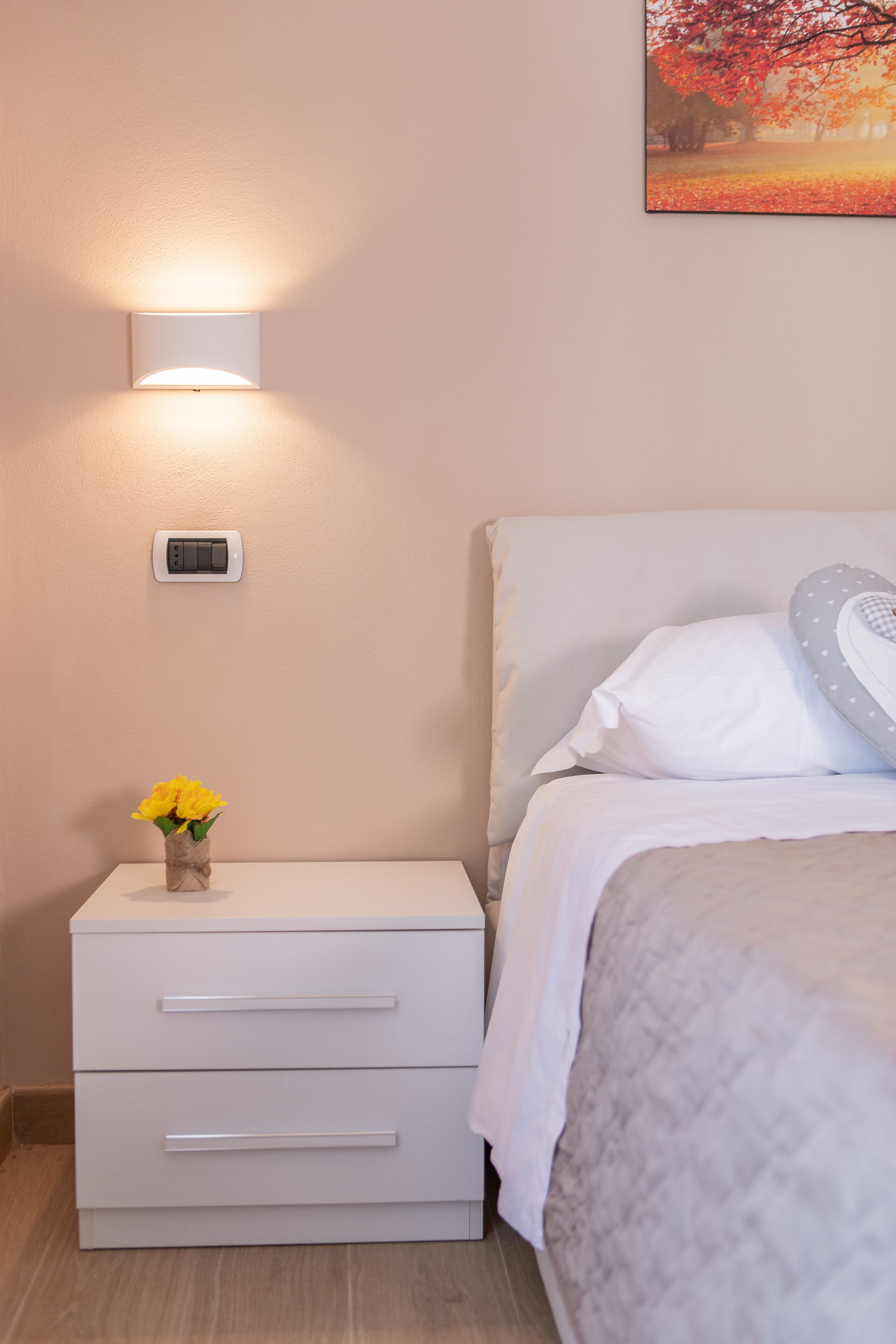 A bedroom with a bed, a bedside table with a lamp, a small bedside table with a vase of flowers, and a thermostat on the wall.