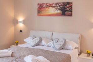 Ein ordentlich bezogenes Bett mit herzförmigen Kissen, ein Nachttisch mit Blumen in einer Vase und ein an der Wand hängendes Bild von einem Baum.
