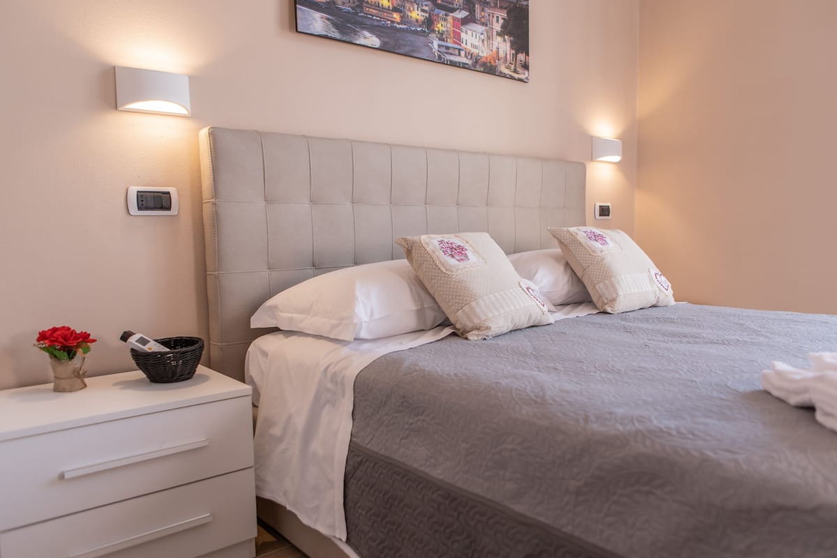 Ein ordentlich bezogenes Bett mit grauem Kopfteil und weißer Bettwäsche, ein Nachttisch mit einer Lampe sowie ein an der Wand befestigtes Thermostat.