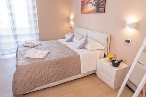 Ein ordentlich bezogenes Bett mit einer beigen Steudecke, Kissen und gefalteten Handtüchern. Ein Nachttisch mit einem Korb, der ein Telefon und eine Vase mit Blumen enthält.