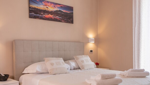 A neatly made bed with white linens and a pink bedspread, a nightstand with a lamp, and a painting of a sunset on the wall. - Le Camere di Teo 2 (La Spezia)