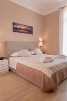 Una cama bien hecha con sábanas blancas y una colcha rosa, una mesita de noche con una lámpara, y un cuadro de un atardecer en la pared.