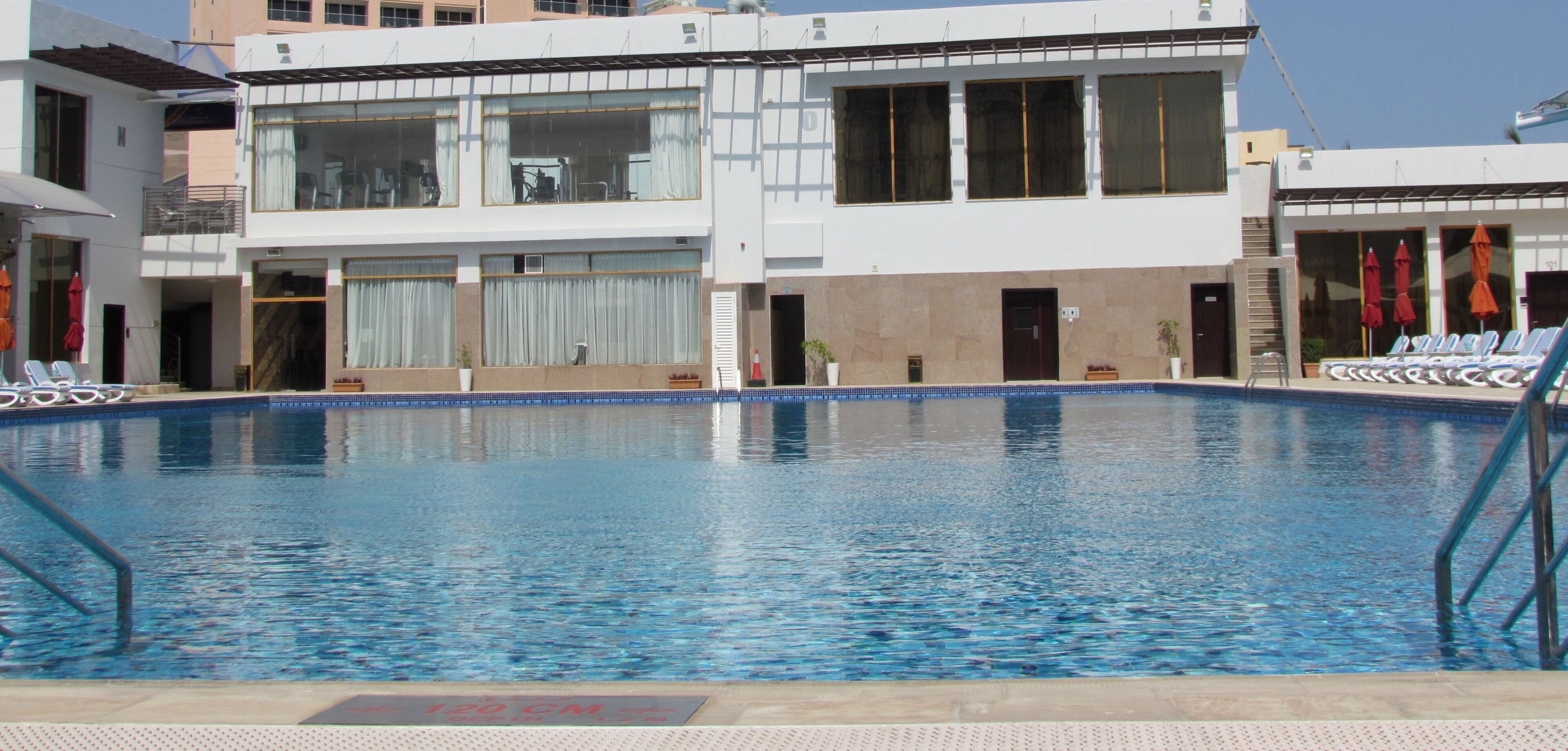 Una piscina con sillas de descanso y sombrillas, rodeada por un edificio con ventanas grandes y un borde de azulejos.