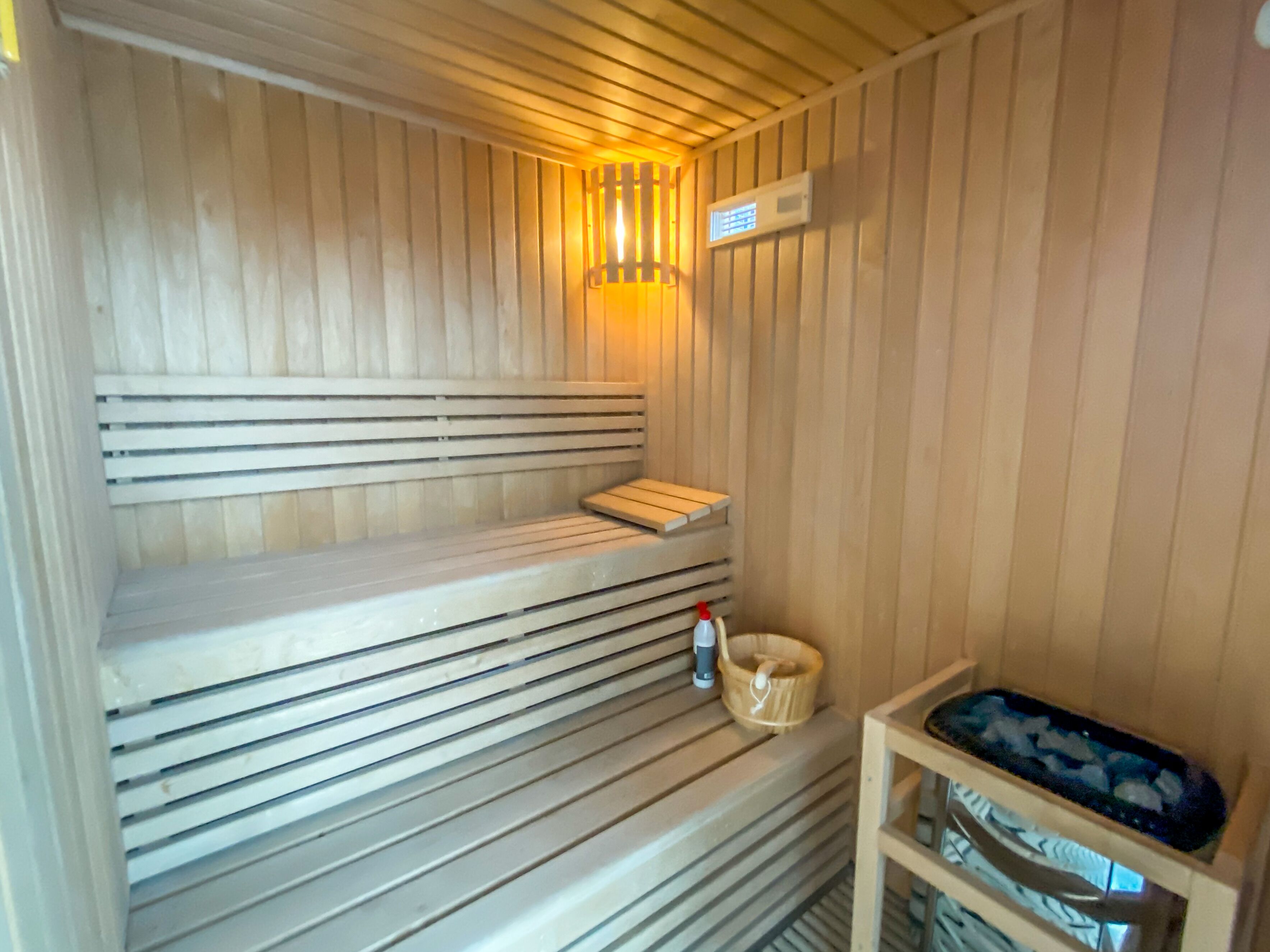Una sauna con bancos de madera, un calentador en la pared, una pequeña mesa de madera con una botella y un recipiente de vidrio a la derecha.