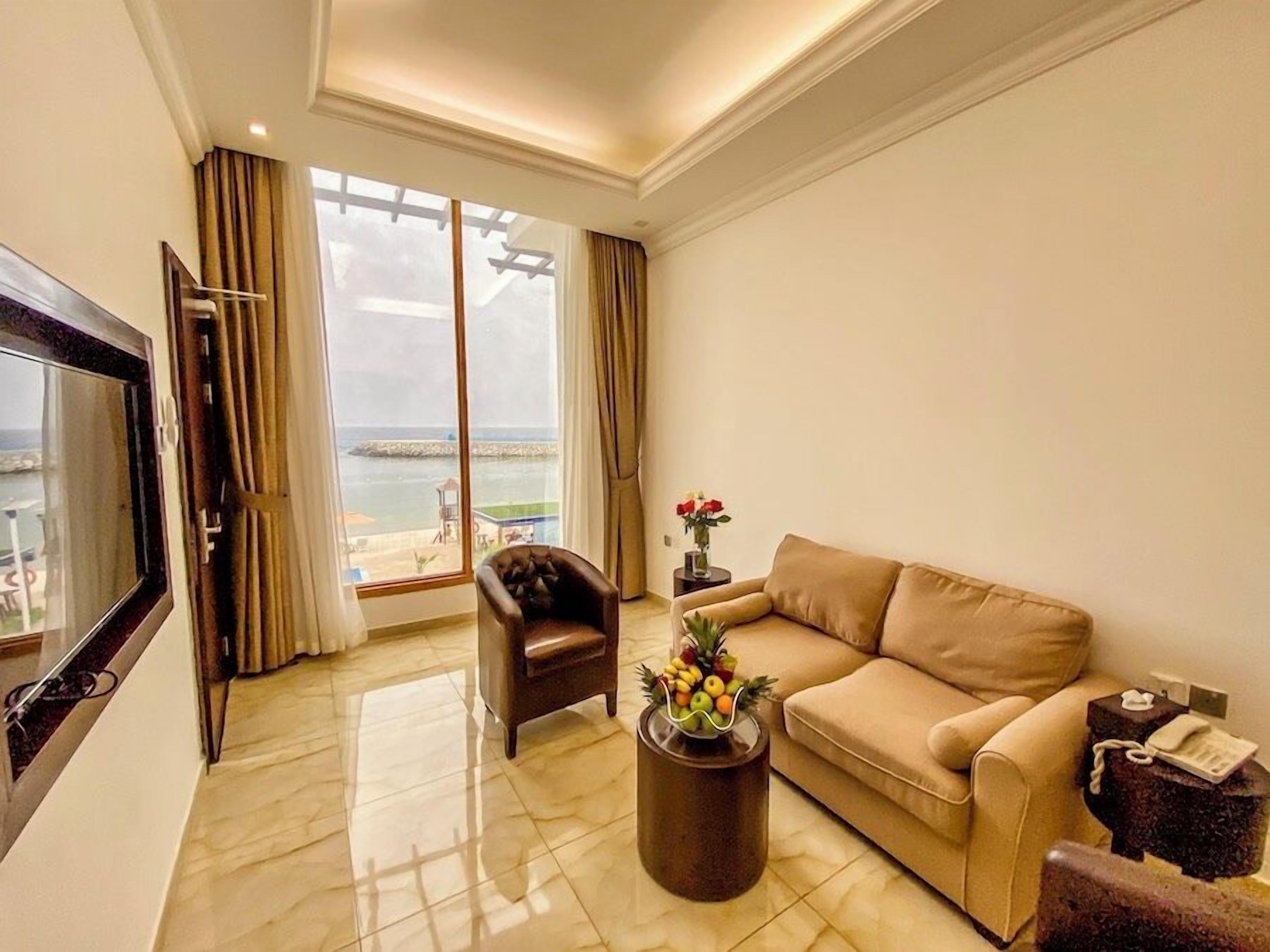 Un salon avec un canapé, un fauteuil et un téléviseur. Une grande fenêtre offre une vue sur la mer.