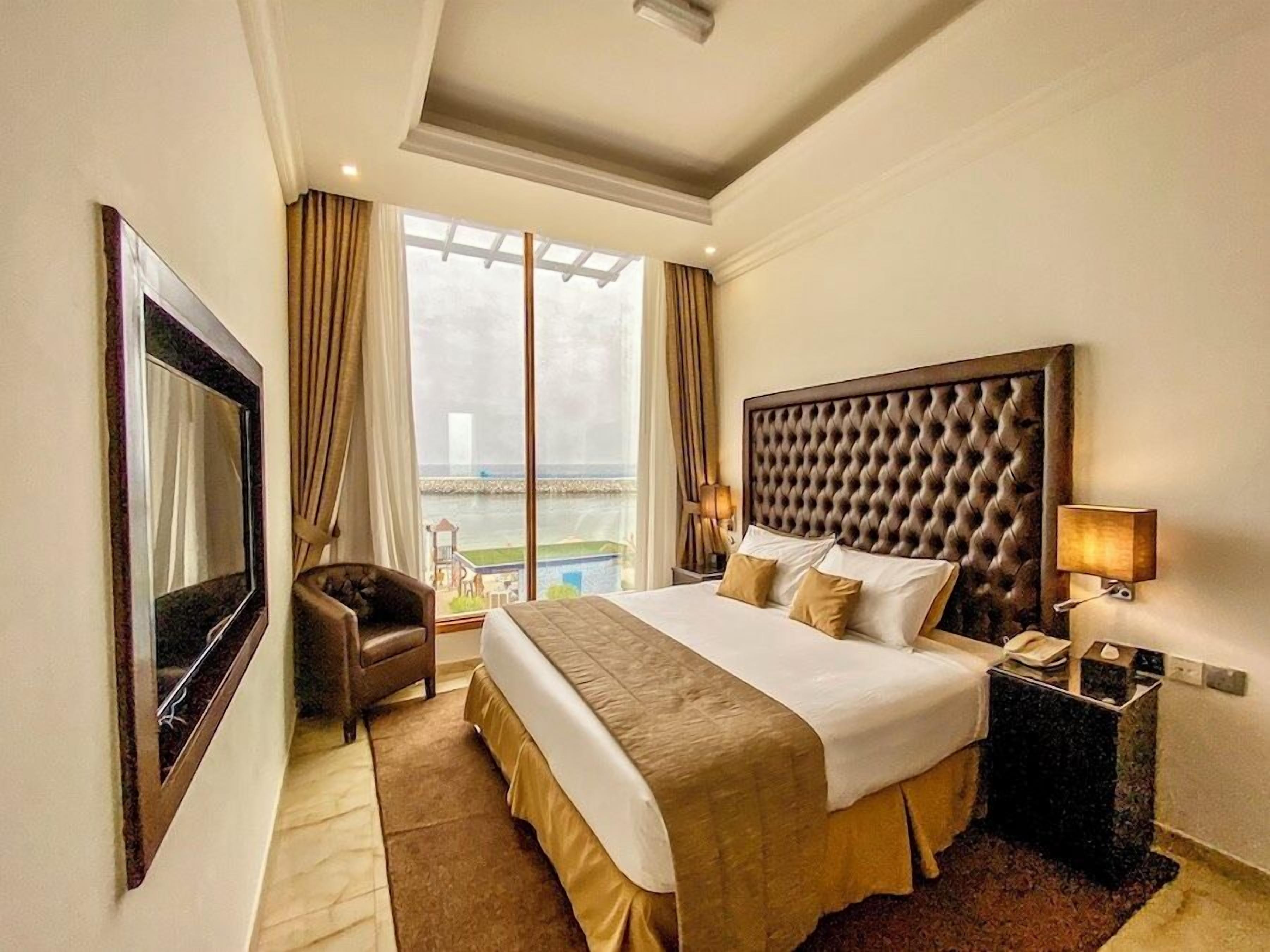Habitación de hotel con una cama grande, una silla, una mesita de noche con una lámpara, un televisor y vistas al mar.