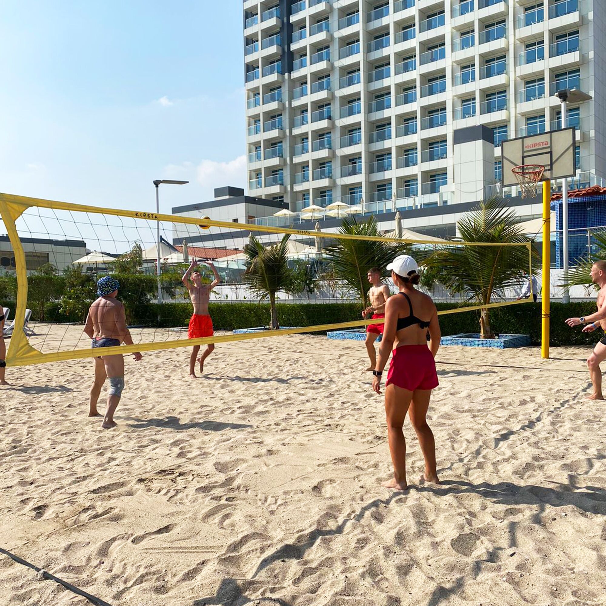 Un match de volleyball de plage se déroule sur un terrain de sable, avec un bâtiment d’hôtel en arrière-plan.