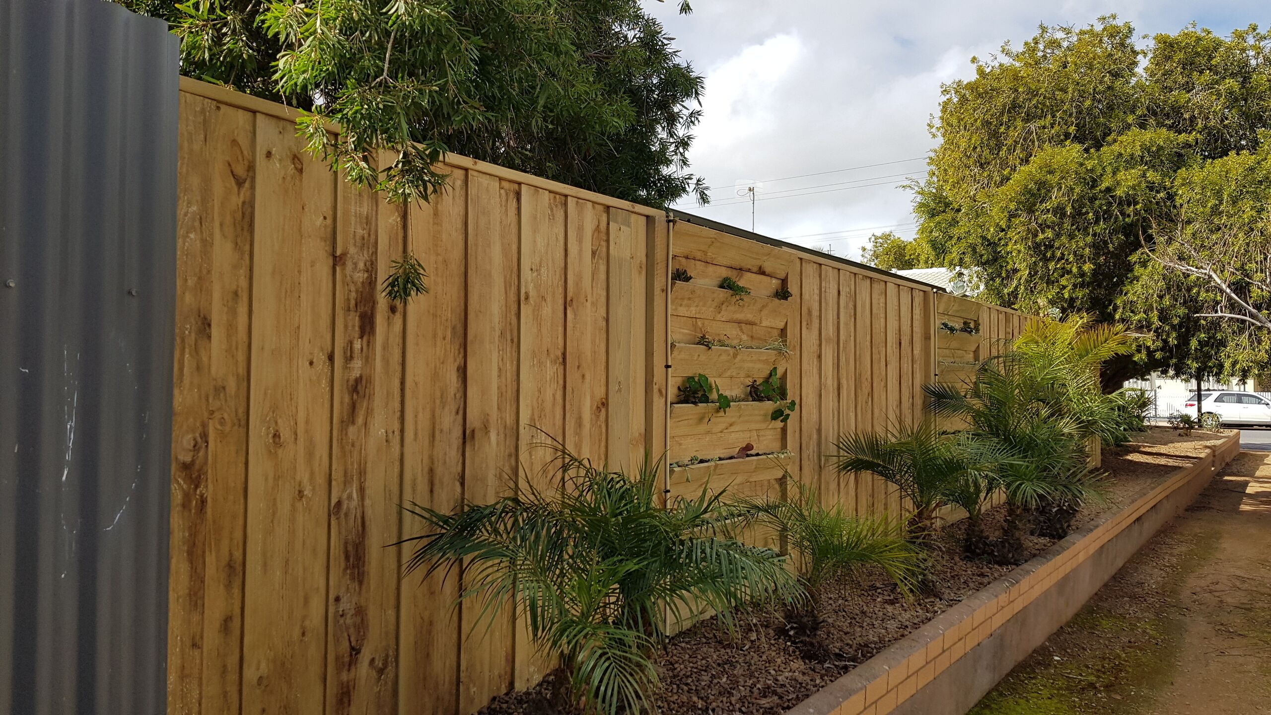 Une clôture en bois avec des lattes verticales et un jardin intégré où sont plantées de petites plantes.