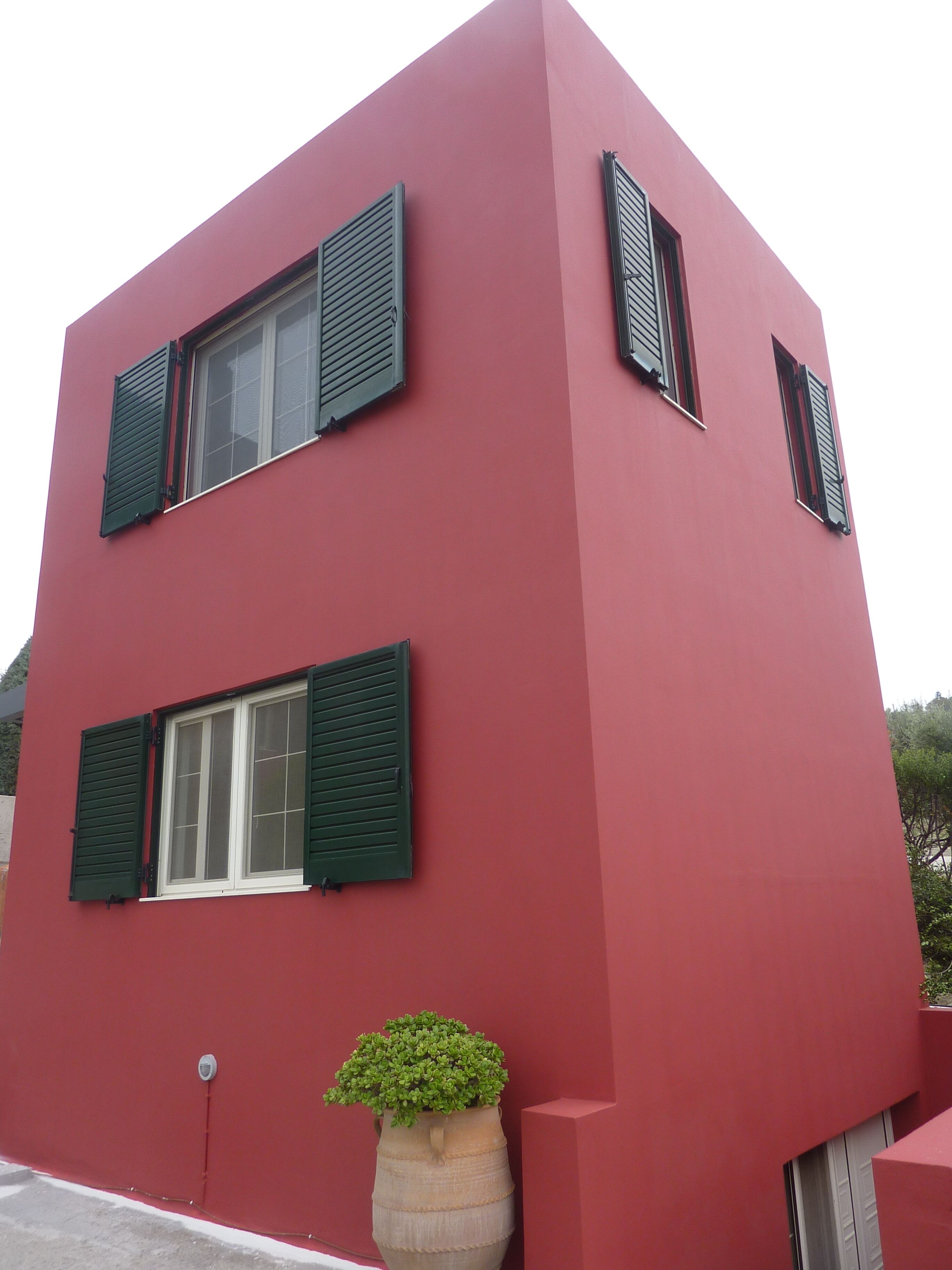 Ein rotes Gebäude mit zwei Fenstern und Fensterläden, einem Blumentopf und einer Sicherheitsvorrichtung.