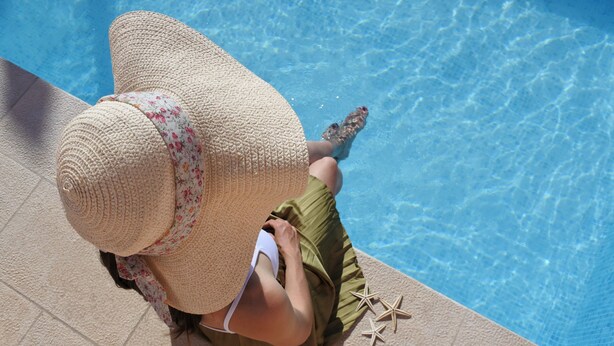 A person sitting by a pool with a large straw hat and a floral pattern, wearing a green skirt and white bikini top.