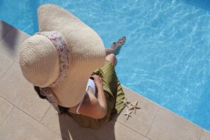 Una persona sentada junto a una piscina, llevando un sombrero de paja grande y estampado floral, con una falda verde y top de bikini blanco.