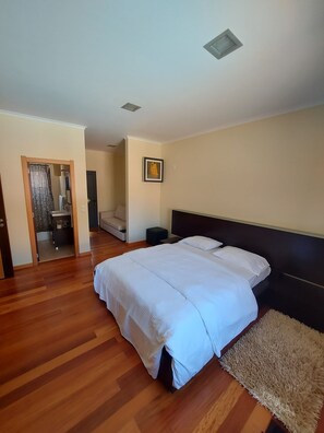 Un dormitorio con una cama grande, piso de madera, cabecera, un cuadro enmarcado en la pared y una puerta que da a otra habitación.