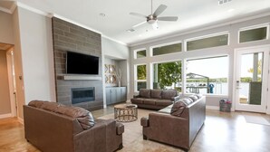 A spacious living room with a fireplace, large windows, and a ceiling fan. - Bud Lane Lakeside | Concierge Services (Kingsland)