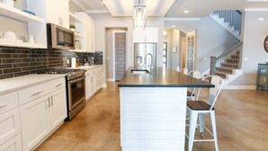 A modern kitchen with white cabinetry, a black tiled backsplash, stainless steel appliances, and a central island with bar stools. - Bud Lane Lakeside | Concierge Services (Kingsland)
