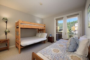 Une chambre avec un lit superposé, un bureau, une table de chevet, une fenêtre donnant sur des arbres et une couvre-lit à motifs.