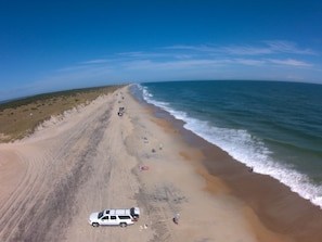 Una playa con un vehículo blanco estacionado en la arena, un cielo despejado y el océano.