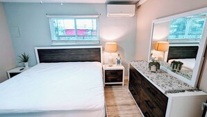 A bedroom with a bed, bedside table, lamp, dresser, mirror, and window. - 1 BR Suite/1st Floor / East Side of Beach Block (Ocean City)