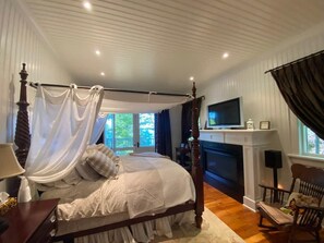 Una cama con dosel, chimenea, televisor y una silla en una habitación con suelo de madera y paredes blancas.