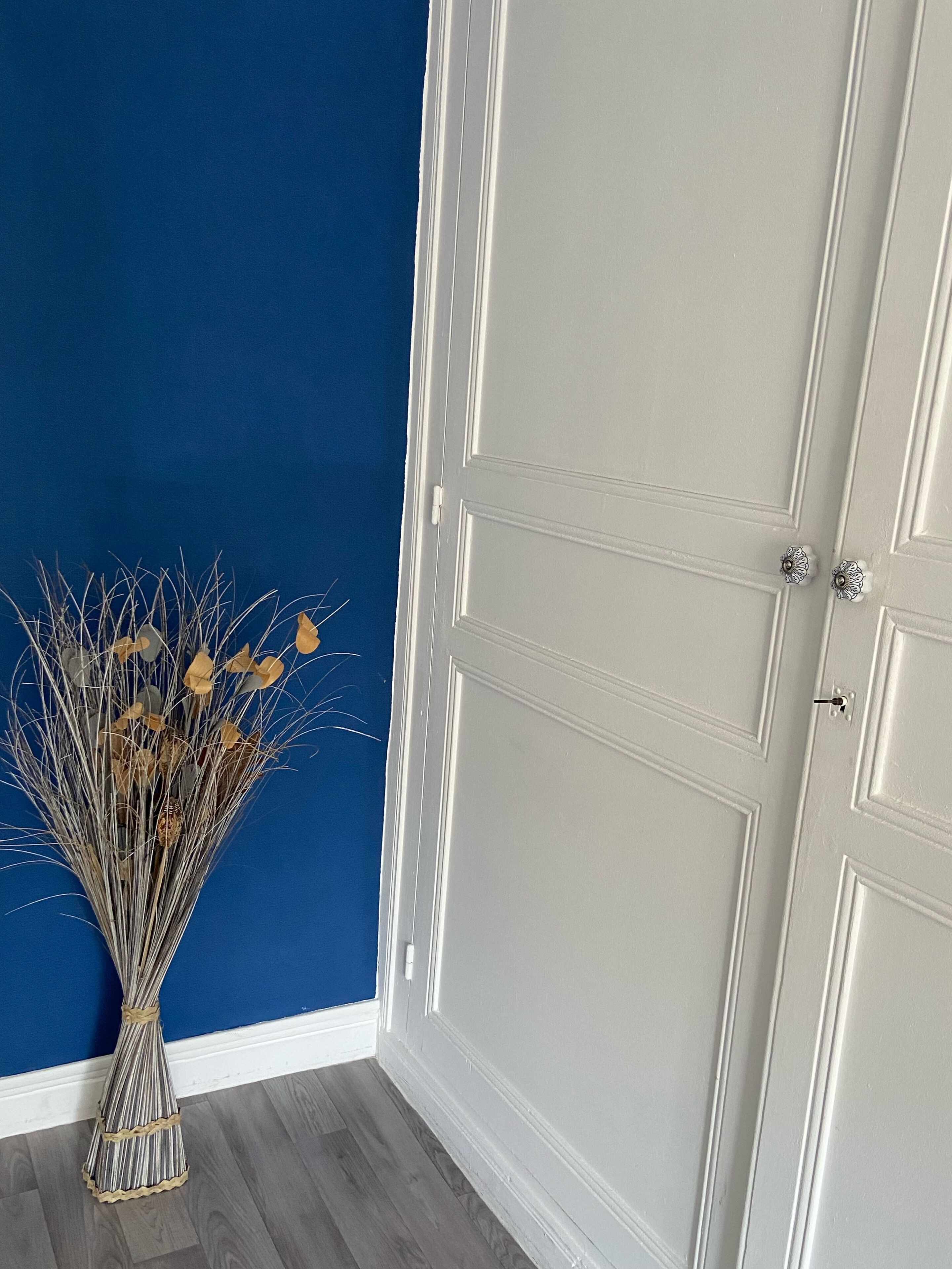 A room with a blue wall, a white door, and a vase of dried flowers.