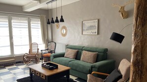 Ein Wohnzimmer mit einem grünen Sofa, einem Holztisch und einem karierten Boden. Es gibt zwei Sessel, eine Lampe und einen wandmontierten Spiegel.