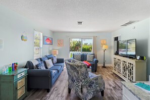 A living room with a sofa, armchair, TV, and a view of greenery outside.