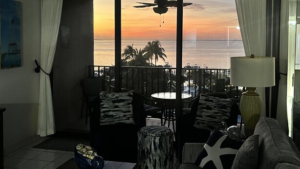 Un balcon offrant une vue sur le coucher de soleil sur l’océan, aménagé avec des chaises et une table, et une lampe placée à droite.