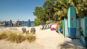 Une plage avec des chaises longues, des parasols et une planche de surf appuyée contre une clôture.