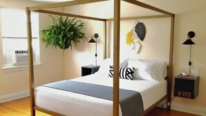 A four-poster bed with a white and gray bedspread, a green plant, and a wall-mounted lamp.