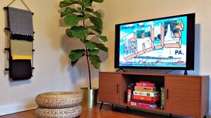 A living room with a television displaying a 'Greetings from Paris' program, a wooden cabinet with board games, a large plant, and woven poufs.