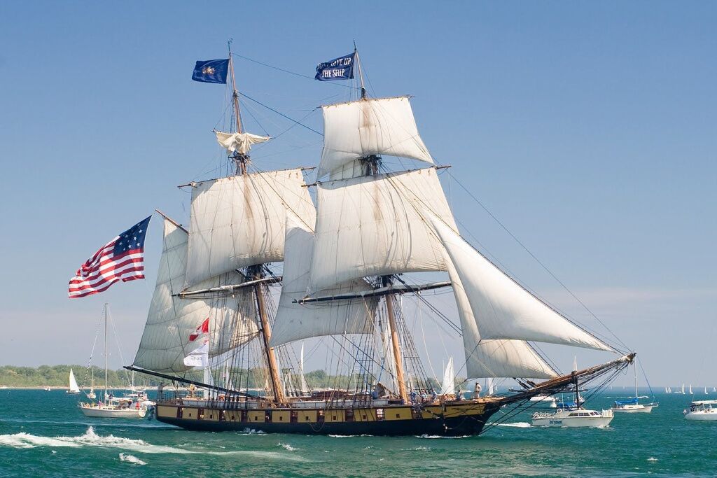 A large sailing ship with multiple masts and sails, anchored in a marina with other boats.