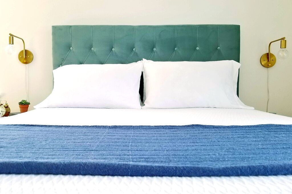 A bed with a teal upholstered headboard, white pillows, and a blue blanket. There are two wall-mounted lamps with round bases and exposed bulbs.