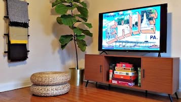 Una sala de estar con una televisión mostrando un programa 'Greetings from Paris', un mueble de madera con juegos de mesa, una planta grande y otomanas tejidas.
