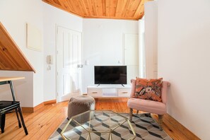 A modern living room with a wooden ceiling, a flat-screen TV on a white cabinet, a pink armchair, and a glass coffee table. - Sun-Drenched Alfama Nest w/ Tagus River Views (Lisboa)