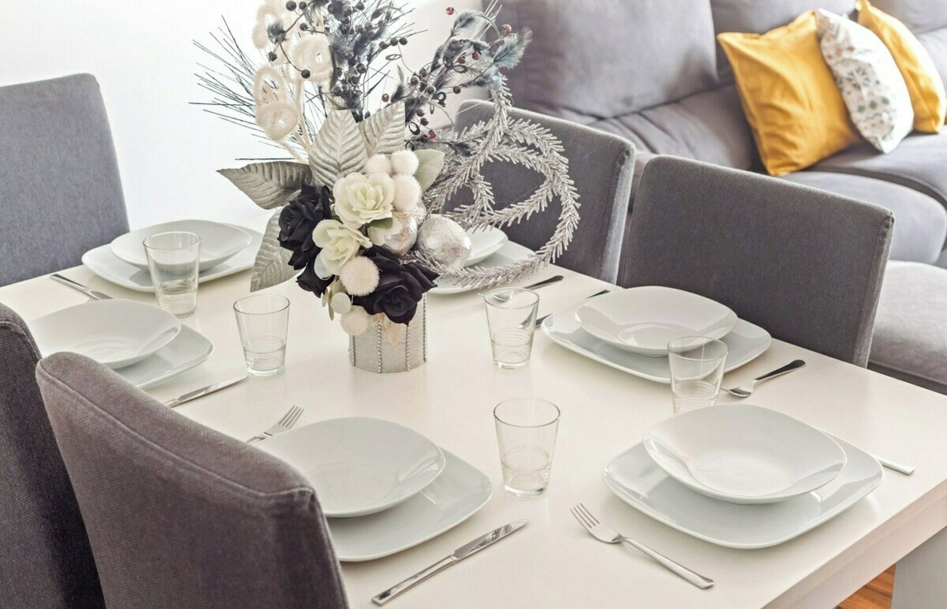Mesa de jantar posta com pratos brancos, copos e talheres, com um arranjo floral decorativo no centro.