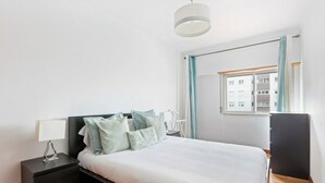 A bedroom with a bed, a bedside table, a lamp, a window with curtains, and a view of buildings outside. - Bright & Spacious Three-Bed Apartment in Lisbon (Lisboa)