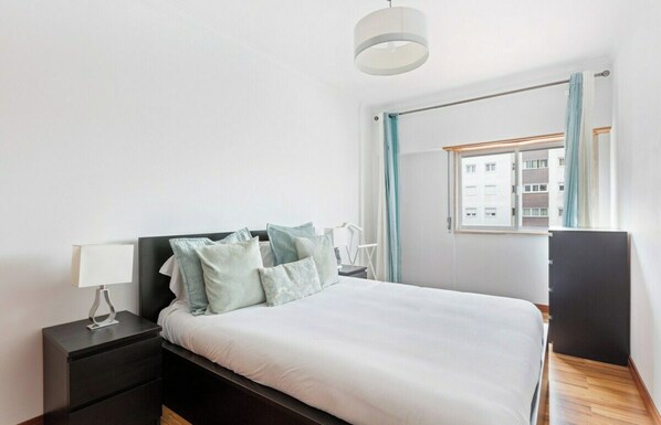 Une chambre à coucher avec un lit, une table de chevet, une lampe, une fenêtre avec des rideaux et une vue sur les bâtiments à l’extérieur.