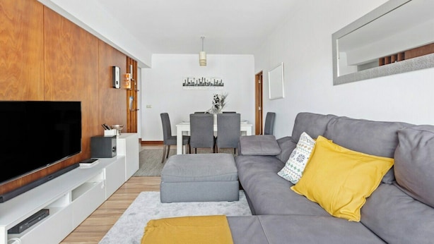A modern living room with a flat-screen TV, grey sofa, ottoman, and a dining area with chairs and a table. - Bright & Spacious Three-Bed Apartment in Lisbon (Lisboa)