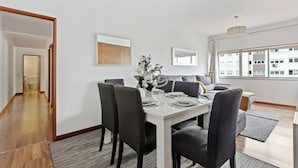 A modern dining area with a white table, black chairs, and a grey sofa. There is a window with a view of buildings and a door leading to another room. - Bright & Spacious Three-Bed Apartment in Lisbon (Lisboa)