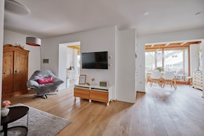 Un salon moderne avec un plancher en bois, un fauteuil gris, un téléviseur fixé au mur et un coin repas comprenant une table et des chaises.