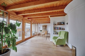 A spacious room with wooden beams, a dining area, a green armchair, and a bookshelf.