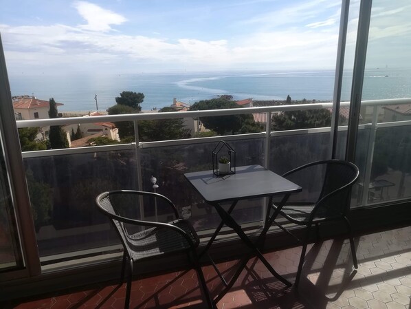 Varanda com duas cadeiras e uma mesa, oferecendo vista para a costa, com edifícios e árvores.